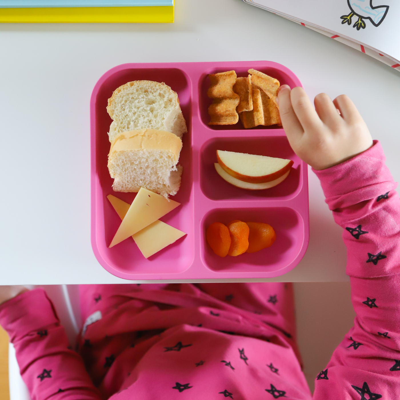 a girl eating her lunch our of a bobo&boo divided plate whihc is bread rolls, cheese, crackers, apples & dried apricot