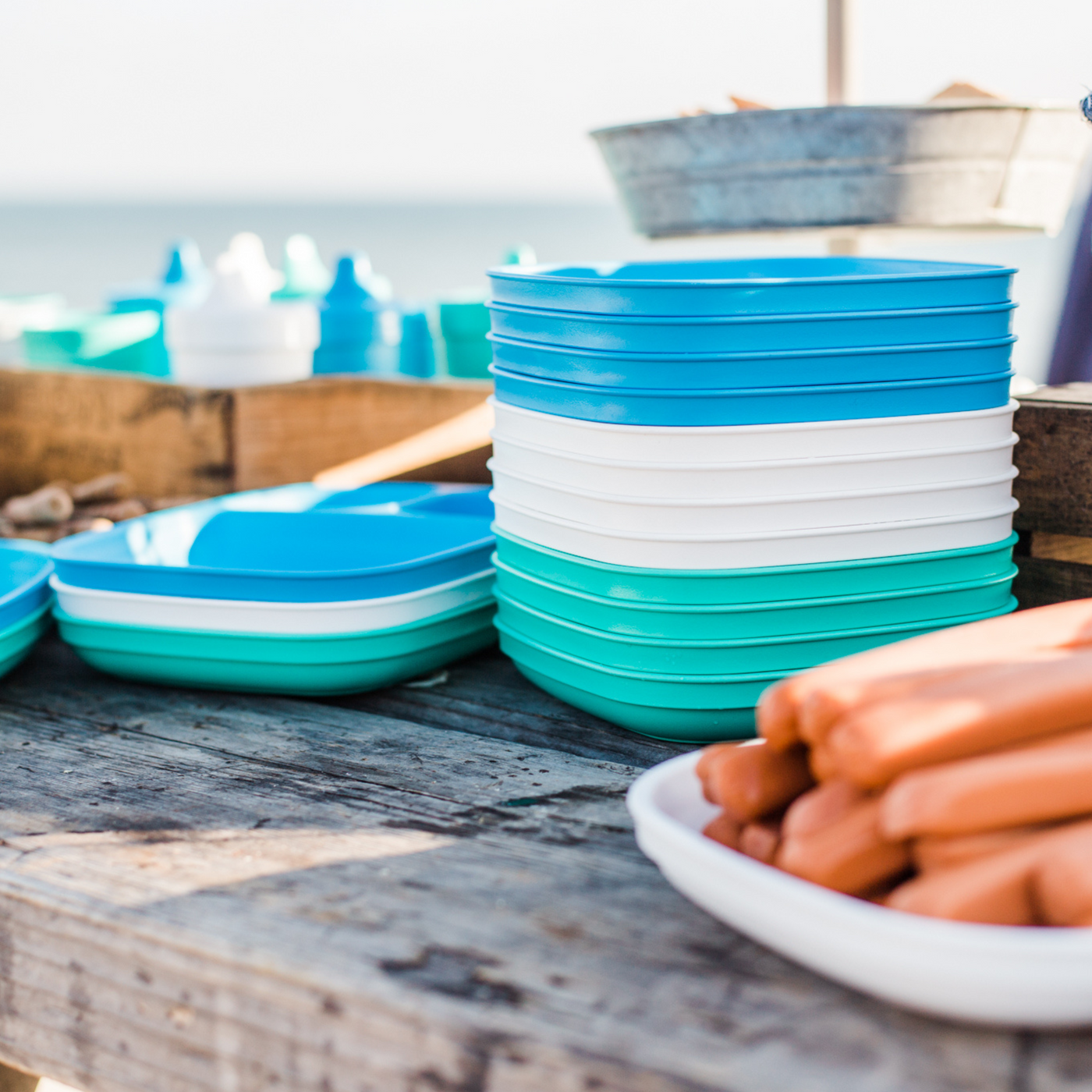 Stacks of blue and white plastic bowls on a wooden surface with hot dogs in the foreground. RePlay Recycled Divided Plate - Teal
