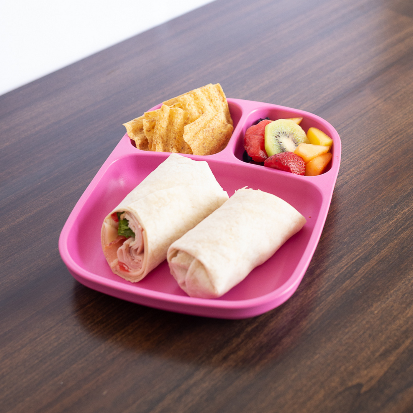 Pink divided tray with wraps, chips, and fruit on a wooden surface. RePlay Recycled Divided Tray Plate - Pink
